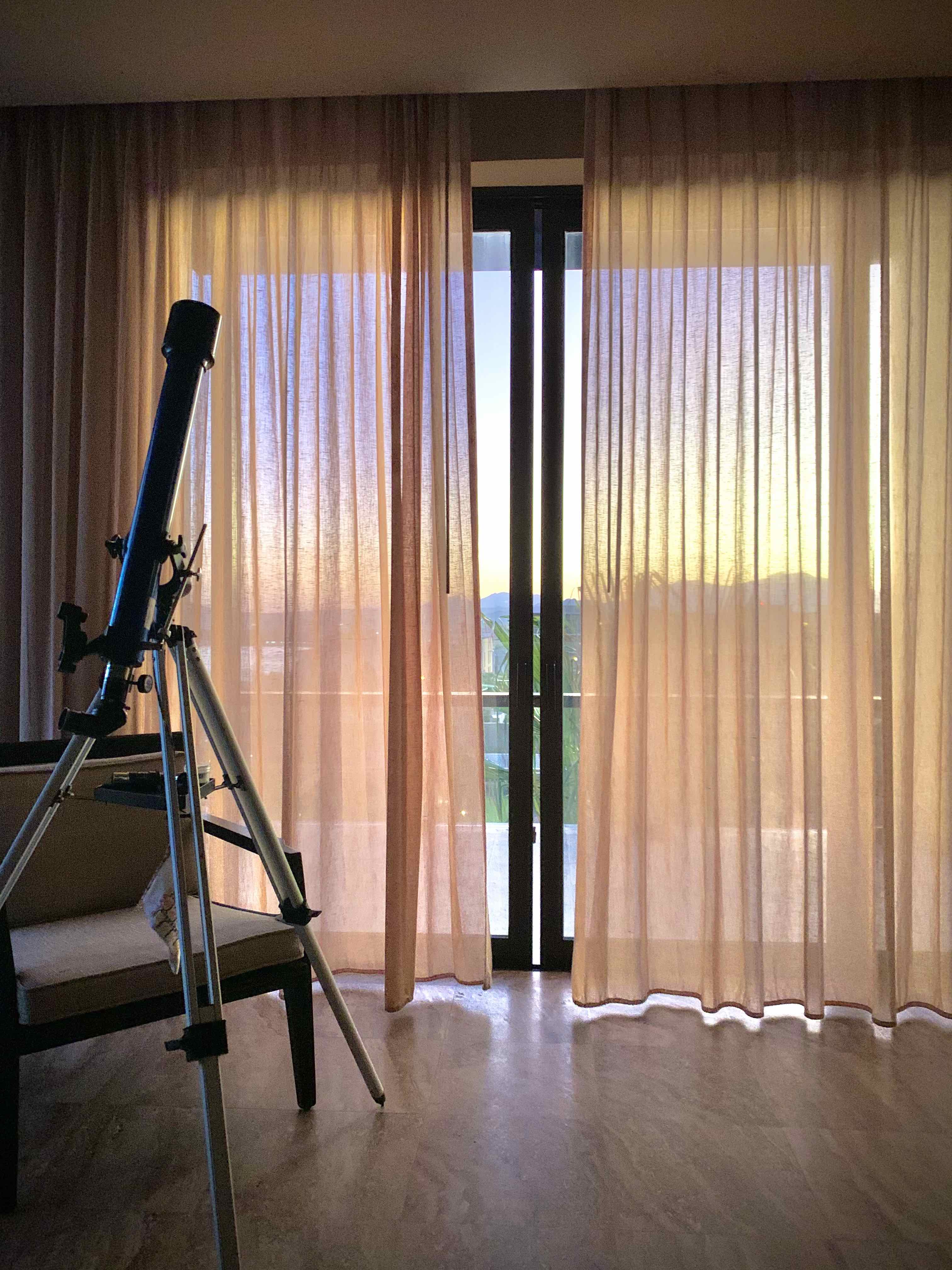 A telescope inside of a room faces the ceiling. Behind it is the draped floor-to-ceiling windows to a balcony featuring a sunset in the distance.